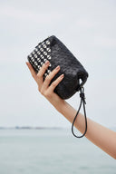 Black woven handbag on a clear pedestal with a yellow object and green sculpture against a blue sky and water background.